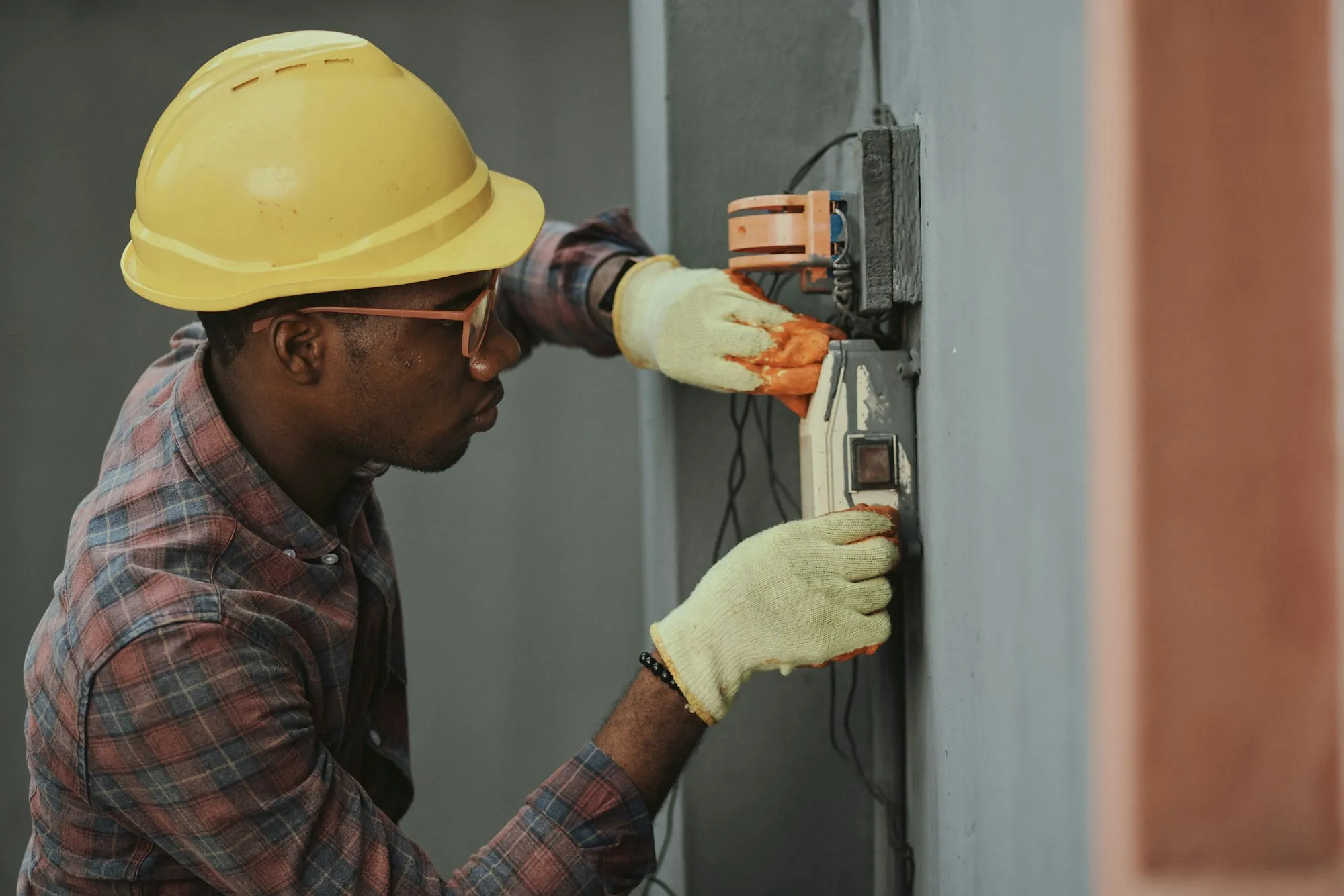 Electrician in a hard hat and gloves working at a wall-mounted electrical box